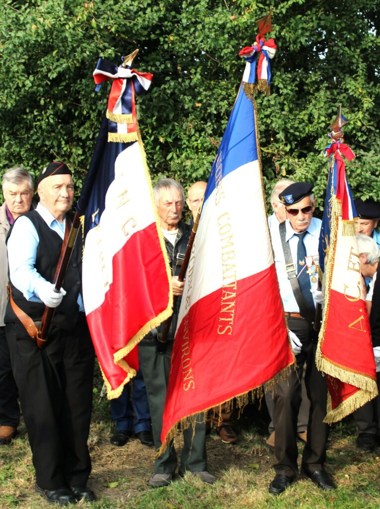 Trois porte-drapeau de Union nationale des combattants de Chaumont-Porcien (Ardennes), monsieur Michel Mauroy de La Romagne (Ardennes) avec son clairon, stèle des fusillés de Wadimont (Ardennes) avec un drapeau du comité de Chaumont-Porcien (Ardennes) du Souvenir français, une croix de Lorraine et d'autres symboles de la Résistance française. Prise de vue effectuée le vendredi 30 août 2024 à Wadimont (Ardennes). Crédits photographiques : © 2020 laromagne.info par Marie-Noëlle ESTIEZ BONHOMME.