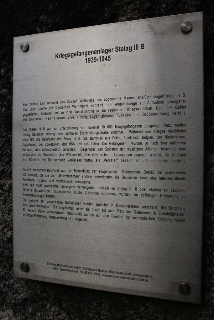 « C'est ici que se trouvait pendant la Seconde Guerre mondiale le Stalag III-B. Ce camp servait à la Wehrmacht pendant ses guerres d'agression pour accueillir les soldats ennemis faits prisonniers et les affecter à l'économie de guerre régionale. Environ soixante-dix camps de même fonction et de même taille étaient répartis sur le territoire du Reich allemand.
Le Stalag III-B était conçu pour accueillir jusqu'à 10 000 prisonniers de guerre. À cette fin, quarante baraques ont été construites le long d'une route centrale. Pendant la guerre, environ 100 000 prisonniers sont passés par le Stalag III-B. Ils venaient de Pologne, de France, de Belgique, des Pays-Bas, de Yougoslavie, d'Union soviétique, des États-Unis et d'Italie. Les prisonniers étaient traités de manière très différente selon leur origine nationale. Les principes du droit international étaient largement respectés à l'égard des soldats des Alliés occidentaux. Les prisonniers italiens, en revanche, leur pays ayant quitté l'alliance avec l'Allemagne, étaient qualifiés de « traîtres » et traités plus durement.
Le traitement réservé aux prisonniers soviétiques était absolument inhumain. Conformément à l'idéologie raciste nazie qui les déclarait « sous-humains », les Allemands leur refusaient une alimentation suffisante, l'hygiène et les soins médicaux.
Plus de 4 000 prisonniers soviétiques sont ainsi morts de faim ou d'épidémies dans le Stalag III-B. D'autres soldats de l'Armée rouge, en particulier ceux de confession juive, ont été transférés vers des camps de concentration pour y être immédiatement assassinés.
Les ossements des prisonniers soviétiques ont d'abord été enterrés dans des fosses communes. Transférés lors de la construction de l'usine sidérurgique en 1951, ils reposent aujourd'hui sur la place du Mémorial à Eisenhüttenstadt. 80 autres victimes de différentes nationalités ont été inhumées dans le cimetière de la paroisse évangélique dans le quartier de Fürstenberg, Kastanienstraße 15 b. »
Photographie en couleurs, prise de vue effectuée le dimanche 27 octobre 2024. Crédits photographiques : © 2020 laromagne.info par Marie-Noëlle ESTIEZ BONHOMME.