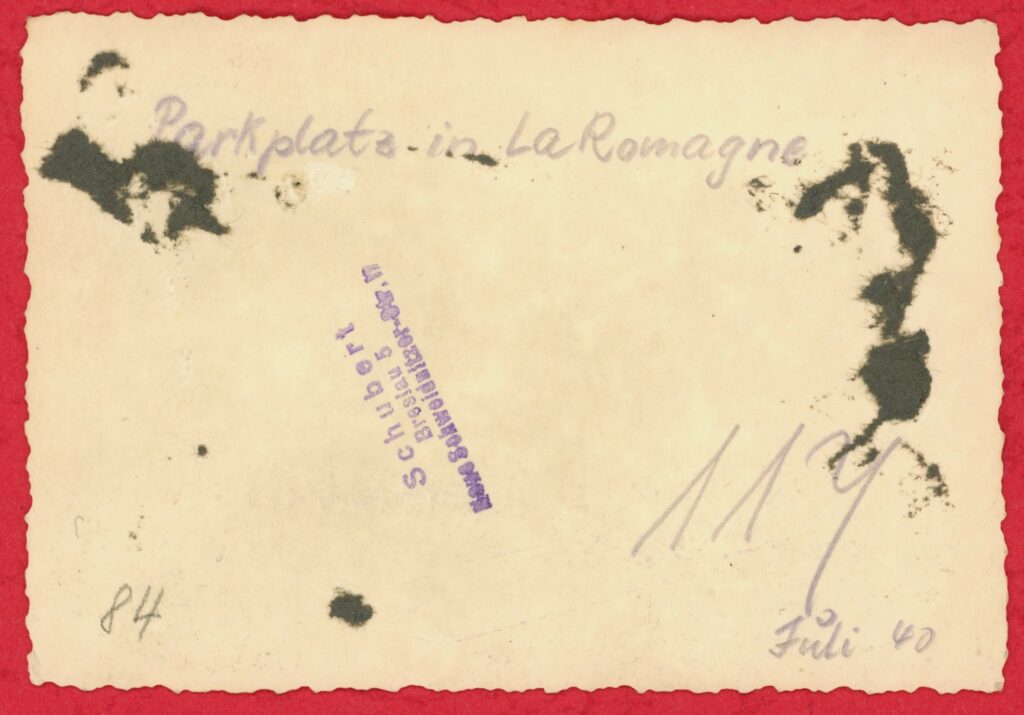 Photographie ancienne, tirage argentique en noir et blanc viré au sépia, sur papier à bords dentelés. Toute reproduction, totale ou partielle, est strictement interdite sans autorisation expresse de l’auteure. Au verso, une écriture manuscrite à l’encre violette indique : « Parkplatz in La Romagne » (« stationnement [de véhicules] à La Romagne »), « Juli 40 » (« juillet 1940 ») et « 104 » [numérotation incertaine]. Un tampon, également à l’encre violette, porte la mention : « Schubert Breslau 5 Neue-Schweidnitzer-Str.17 ». (Exemplaire conservé dans la collection personnelle de l’auteure.)
