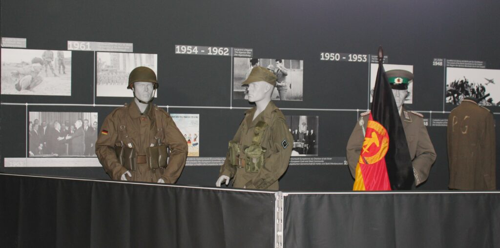 Uniformes allemands accompagnés du drapeau de la République Démocratique Allemande : tricolore horizontal noir-rouge-or, chargé en son centre des armoiries de la RDA (marteau et compas entourés d’une gerbe de blé ceinte d’un ruban aux mêmes couleurs). Prise de vue effectuée le lundi 5 mai 2025 au musée Guerre et Paix en Ardennes (Novion-Porcien, Ardennes). Crédits photographiques : © 2020 laromagne.info par Marie-Noëlle ESTIEZ BONHOMME.