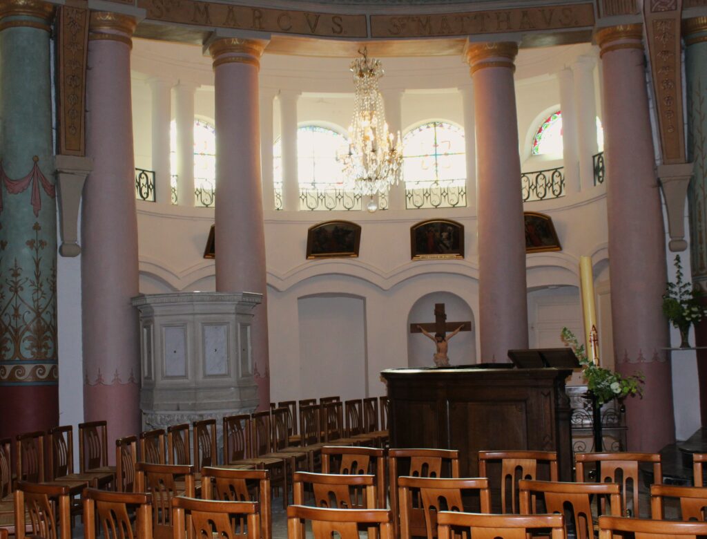 Au fond du chœur de l'église Saint-Didier d'Asfeld (Ardennes) se dresse le maître-autel, adossé, conçu pour la célébration selon le rite romain antérieur à la réforme liturgique. Au premier plan, l'autel de célébration, disposé face au peuple conformément aux orientations issues du concile Vatican II, est orné d'un devant d'autel sculpté en bois réalisé par Didier Romagny. L'éclairage est assuré par des lustres à pampilles, qui contribuent à la solennité et à l'élégance de l'édifice. Photographies en couleurs, prises de vue réalisées le samedi 25 avril 2026. Crédits photographiques : © 2020 laromagne.info par Marie-Noëlle ESTIEZ BONHOMME.