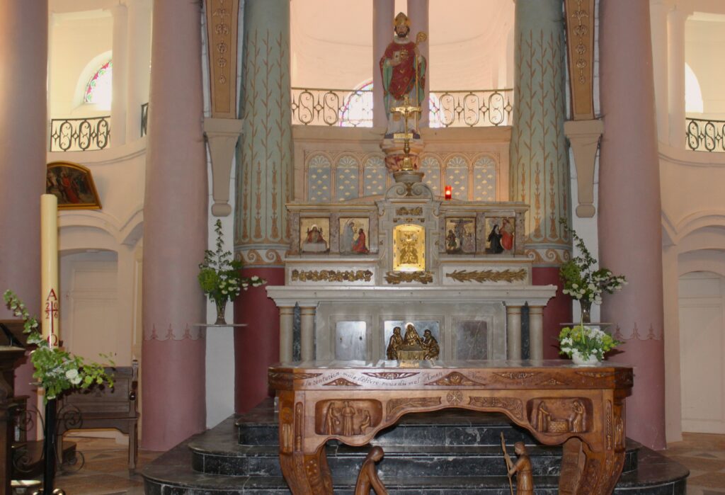 Au fond du chœur de l'église Saint-Didier d'Asfeld (Ardennes) se dresse le maître-autel, adossé, conçu pour la célébration selon le rite romain antérieur à la réforme liturgique. Au premier plan, l'autel de célébration, disposé face au peuple conformément aux orientations issues du concile Vatican II, est orné d'un devant d'autel sculpté en bois réalisé par Didier Romagny. L'éclairage est assuré par des lustres à pampilles, qui contribuent à la solennité et à l'élégance de l'édifice. Photographies en couleurs, prises de vue réalisées le samedi 25 avril 2026. Crédits photographiques : © 2020 laromagne.info par Marie-Noëlle ESTIEZ BONHOMME.