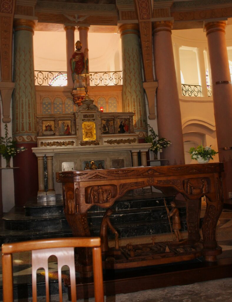 Au fond du chœur de l'église Saint-Didier d'Asfeld (Ardennes) se dresse le maître-autel, adossé, conçu pour la célébration selon le rite romain antérieur à la réforme liturgique. Au premier plan, l'autel de célébration, disposé face au peuple conformément aux orientations issues du concile Vatican II, est orné d'un devant d'autel sculpté en bois réalisé par Didier Romagny. L'éclairage est assuré par des lustres à pampilles, qui contribuent à la solennité et à l'élégance de l'édifice. Photographies en couleurs, prises de vue réalisées le samedi 25 avril 2026. Crédits photographiques : © 2020 laromagne.info par Marie-Noëlle ESTIEZ BONHOMME.