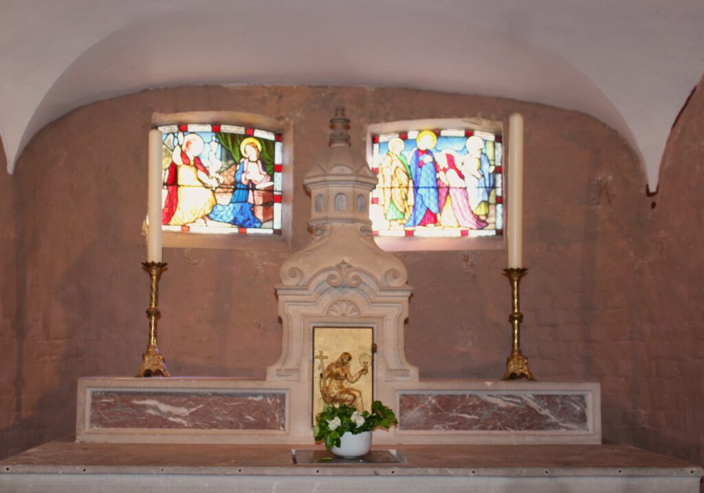 Au fond du chœur de l'église Saint-Didier d'Asfeld (Ardennes) se dresse le maître-autel, adossé, conçu pour la célébration selon le rite romain antérieur à la réforme liturgique. Au premier plan, l'autel de célébration, disposé face au peuple conformément aux orientations issues du concile Vatican II, est orné d'un devant d'autel sculpté en bois réalisé par Didier Romagny. L'éclairage est assuré par des lustres à pampilles, qui contribuent à la solennité et à l'élégance de l'édifice. Photographies en couleurs, prises de vue réalisées le samedi 25 avril 2026. Crédits photographiques : © 2020 laromagne.info par Marie-Noëlle ESTIEZ BONHOMME.