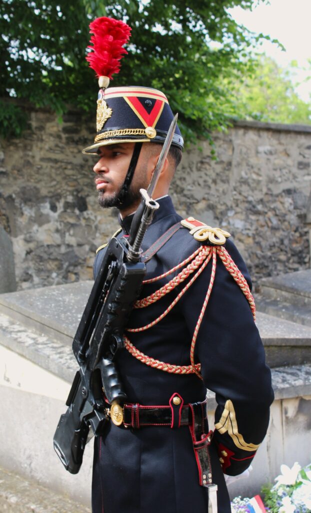Ce garde républicain porte très probablement un fusil d’assaut FAMAS F1, encore utilisé pour les services d’honneur, bien que le HK416F ait remplacé le FAMAS dans les unités opérationnelles. L’acronyme FAMAS signifie « Fusil d’assaut de la Manufacture d’armes de Saint-Étienne ». Photographie en couleurs, prise de vue réalisée le mercredi 20 mai 2026. Crédits photographiques : © 2020 laromagne.info par Marie-Noëlle ESTIEZ BONHOMME.
