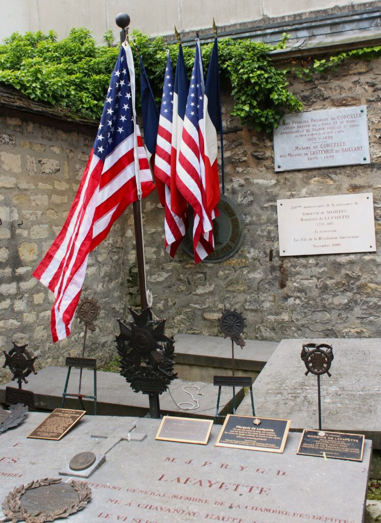 Drapeaux des États-Unis déployés au cimetière de Picpus (Paris, Île-de-France), devant les plaques commémoratives du marquis de La Fayette et des liens historiques franco-américains. Photographie en couleurs. Prise de vue réalisée le mercredi 20 mai 2026. Crédits photographiques : © 2020 laromagne.info par Marie-Noëlle ESTIEZ BONHOMME.